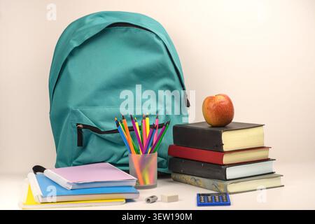 Turquoise school backpack with pencil cup, stack of books, red apple, notebooks, calculator, eraser and sharpener on a clean light background. Stock Photo