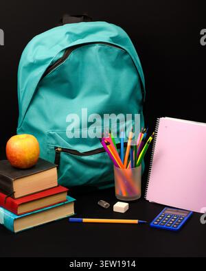 Turquoise backpack with books, apple, colored pencils, notebook, calculator, eraser and sharpener on black background, back to school concept. Stock Photo