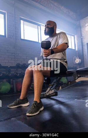 Bearded man wearing gray T-shirt lifting hex dumbbell while sitting on flat bench at gym Stock Photo