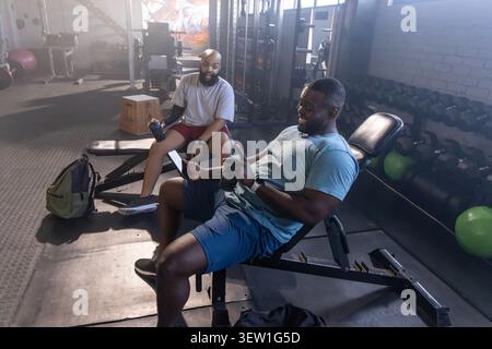 African American men sitting on incline bench at gym wearing shorts holding green shaker and phone Stock Photo