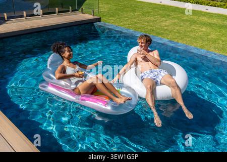 Couple lounging on inflatable floats in backyard pool, holding clear cups, sunlit reflections Stock Photo
