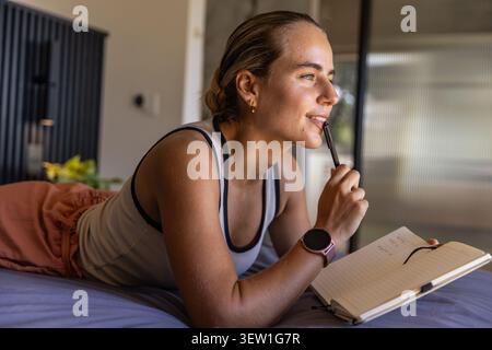 Female lying on stomach on bed, holding pen near mouth with open notebook and pink smartwatch Stock Photo