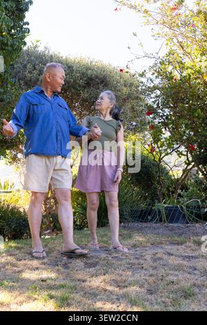 Asian senior couple holding hands, gazing at each other on patchy lawn with potted plants Stock Photo