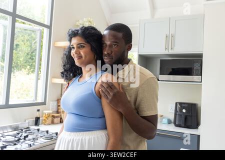 Diverse couple embracing in kitchen by window and cooktop, wearing blue top and beige shirt Stock Photo