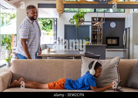 African American dad standing at home, boy lying on sofa wearing headset using tablet, copy space Stock Photo