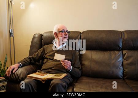 Senior man sitting on a couch, holding an old photograph and photo album, looking away in thought Stock Photo