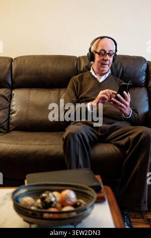 Senior man sitting on a sofa wearing headphones and using a smartphone Stock Photo