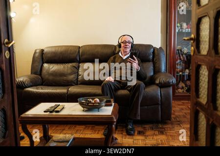 Senior man listening to music on headphones while using smartphone at home Stock Photo