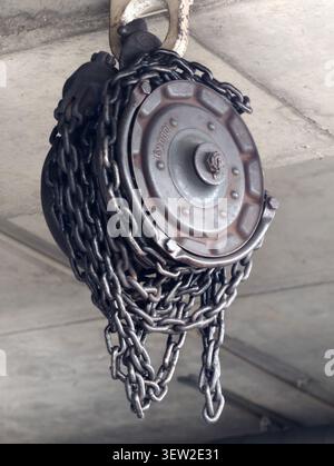 Closeup of industrial chain hoist pulley assembly suspended from concrete ceiling hook, featuring coiled heavy steel chain, rusted stamped pulley cove Stock Photo