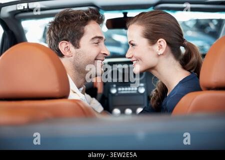 Back, car dealership and couple in convertible for decision, purchase or test drive. Choice, love and satisfaction with happy people in showroom Stock Photo