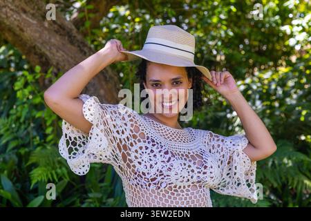 African American woman standing smiling, holding straw hat, wearing lace top in garden by tree Stock Photo