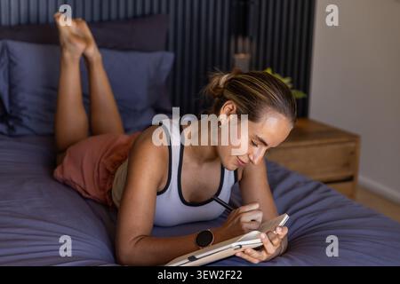 Woman lying on stomach on bed in bedroom, using stylus on tablet, wearing tank top Stock Photo