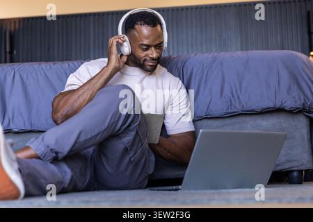 African mature man sitting in bedroom beside bed wearing headphones, holding tablet, using laptop Stock Photo