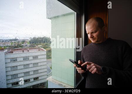 Man texting on smartphone by window with raindrops, experiencing loneliness during a gloomy day Stock Photo