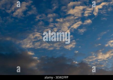 Scattered white and golden altocumulus clouds across a deep blue sky with darker cloud mass along the lower edge Stock Photo