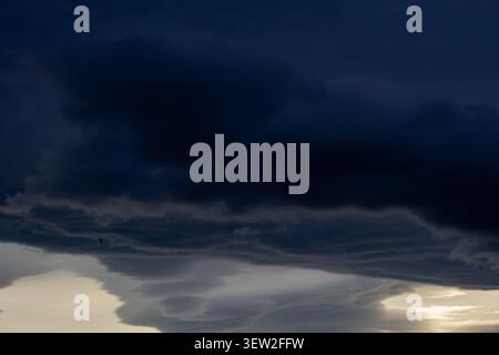 Dramatic stormy sky with dark navy clouds above pale lower cloud formations with a tiny bird in flight and light on the horizon Stock Photo