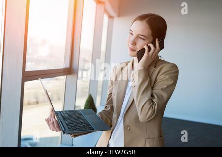 Professional businesswoman multitasking with laptop and smartphone by window Stock Photo