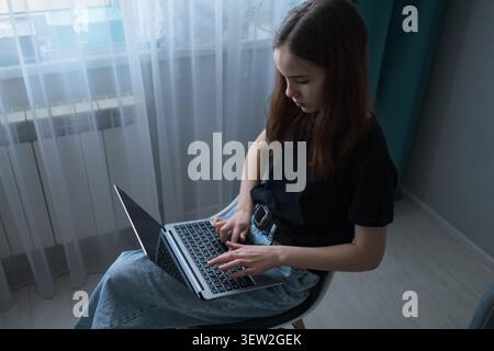 Young woman focusing on her laptop, typing on the keyboard while sitting on a chair, engaged in online learning or remote work activities inside her Stock Photo