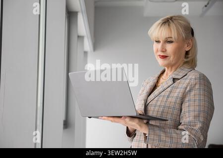 Blonde middle aged businesswoman working with a laptop, standing confidently by a window in a bright corporate environment, embodying focus and Stock Photo