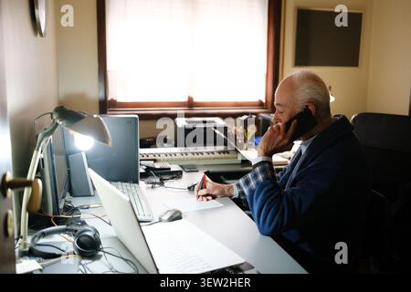 Senior man working from home talking on smartphone and writing notes at his desk Stock Photo