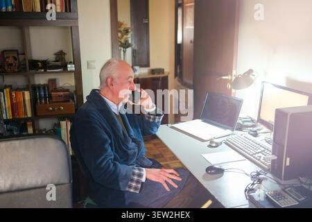 Senior man talking on smartphone while working remotely from his home desk Stock Photo