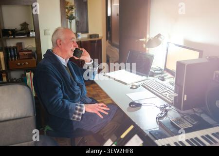 Senior man in bathrobe working remotely from home office speaking on smartphone Stock Photo