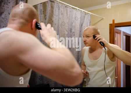 Man grooming his head with an electric shaver, looking at his reflection in the mirror Stock Photo