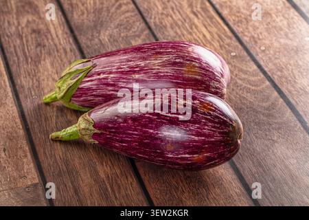 Two glossy striped eggplants with deep purple white streaks rest on dark wood planks. Natural lighting highlights texture sheen. Composition emphasize Stock Photo
