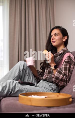 Young woman relaxing on sofa, enjoying a cookie and hot drink in her cozy living room at home. Snacking while watching tv Stock Photo