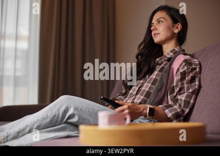 Young woman sitting comfortably on a couch with a remote control, enjoying leisure time and watching television at home Stock Photo