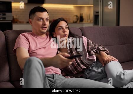 Young couple sitting on a sofa in the living room, looking surprised, amazed, and shocked while watching television Stock Photo
