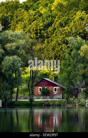 Red lakeside cabin nestles among tall trees with soft reflections in still water dusk tones add warmth while nature surrounds the cozy retreat invitin Stock Photo