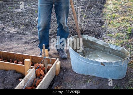 Worker mixing concrete beside wooden formwork. Worker stands beside a metal tub of wet concrete and wooden formwork filled with bricks on an outdoor Stock Photo