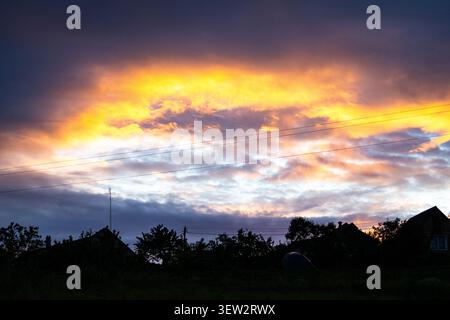A fiery window opens in storm clouds above a rural line of houses. Electric gold pours through churning layers while rooftops rest in silhouette as th Stock Photo