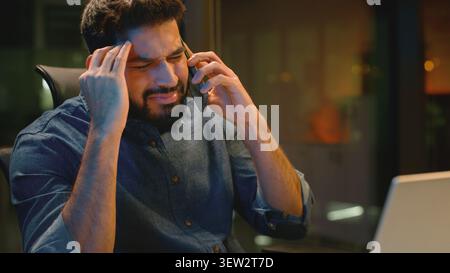 Annoyed furious angry Indian business man arguing phone looking in laptop at night office dissatisfied Arabian businessman male worker executive argue Stock Photo