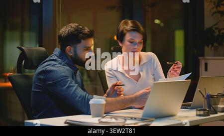 Diversity two business colleagues talking review documents and laptop in dark night office Indian businessman Arabian man talk helping Caucasian woman Stock Photo