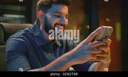 Happy Indian business man browsing mobile phone in evening office at table joyful Arabian businessman smiling scrolling web app Hindu male guy Stock Photo