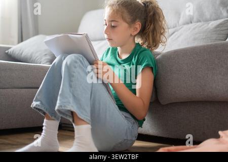 Elementary school girl sitting on the floor in a cozy living room, reading and writing in a notebook Stock Photo