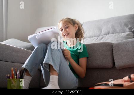 Elementary school girl sitting on the floor in a cozy living room, reading and writing in a notebook Stock Photo