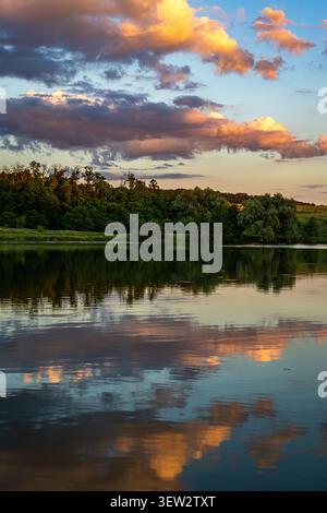Warm clouds drift above a green forest and their reflections shimmer on the placid lake inviting a calm contemplative pause suited to wellness travel Stock Photo