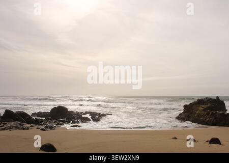 Ocean waves washing over dark rocks on a sandy beach under a cloudy sky Stock Photo