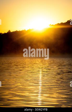 The sun climbs and lays a burning ribbon on the rippled water. Steam fades into the hillside and the lake breathes gold. Silence gathers and the day o Stock Photo