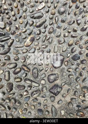 A top-down view of a textured concrete pavement embedded with smooth, rounded grey and brown reflexology pebbles. Best for wallpaper and background. Stock Photo