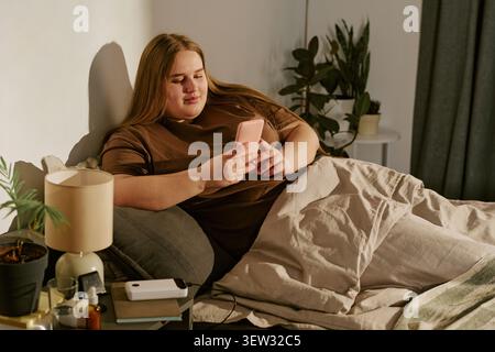 Plus Size Woman Relaxing in Bed While Using Smartphone at Home Stock Photo