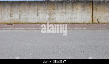 Grunge prefab concrete boundary wall with with weathering marks, stains, moss, and lichens. Sidewalk and street in front. Background for copy space Stock Photo