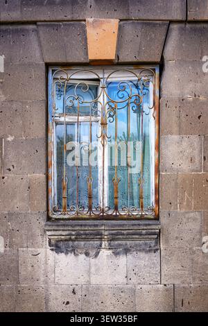 Old stone wall with ornate metal window grate. Window reflects blue sky and clouds. Rusty metal bars add vintage charm. Weathered brickwork shows time Stock Photo