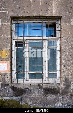 Old window with metal bars set in weathered stone wall. Glass panes show signs of age and wear. Rusty metal grid covers broken window sections. Stone Stock Photo