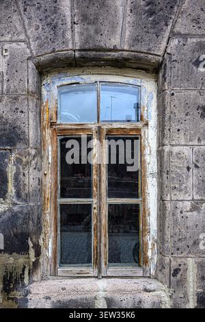Old, weathered window set in stone wall. Wood frame shows decay and rust. Glass panes reflect sky and clouds. Stone blocks around window look worn and Stock Photo