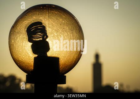 A close-up, backlit shot of a streetlamp with a compact fluorescent bulb inside a scratched, amber-tinted globe, silhouetted against a warm, clear sky Stock Photo
