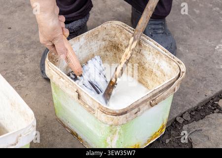 Worker blending liquid plaster or cement mixture in a dirty container using a tool for home improvement projects, highlighting professional masonry sk Stock Photo